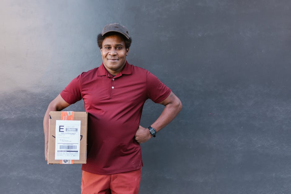 A smiling delivery person with medium brown skin and curly black hair, wearing a maroon polo shirt, matching cap, and black watch, stands against a plain gray wall. He holds a rectangular cardboard pa