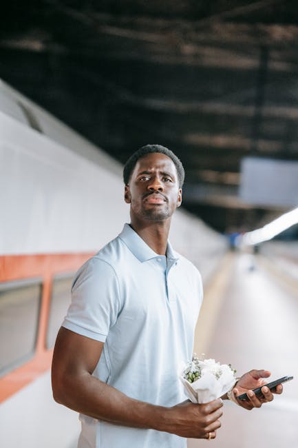 A man with short curly hair, wearing a light blue polo shirt, stands on a train platform holding a bouquet of white flowers wrapped in white paper in one hand and a smartphone in the other. The backgr