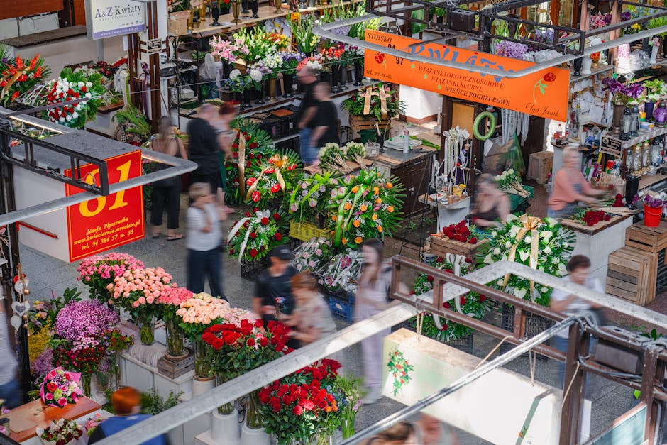 An indoor flower market bustling with activity, featuring vibrant floral arrangements including pink chrysanthemums, red roses, purple asters, and white lilies in various vases and baskets. The flower