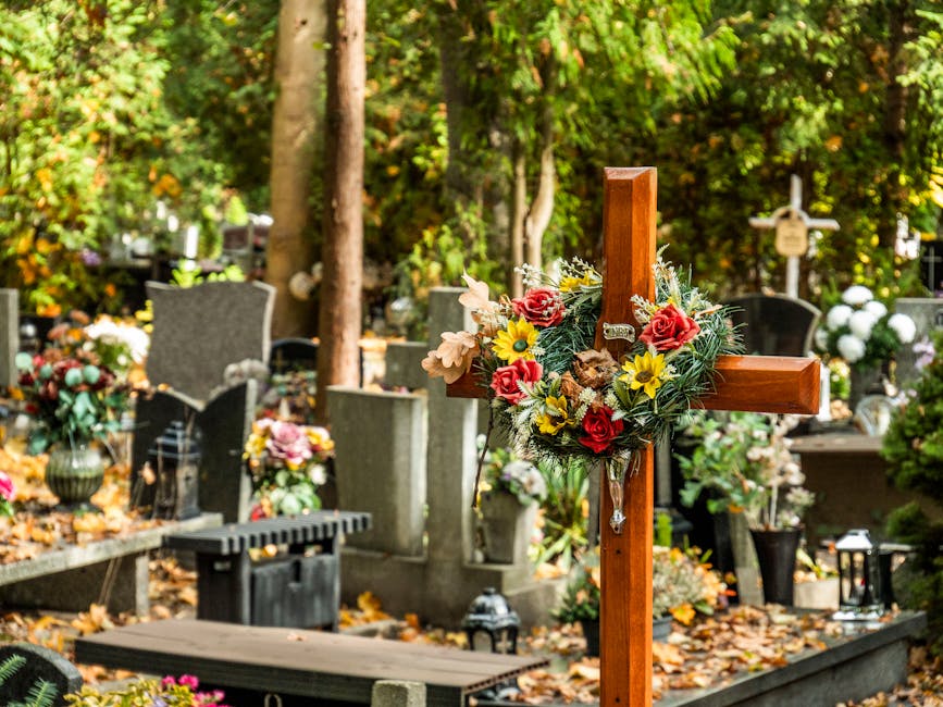 A wooden funeral cross adorned with a floral arrangement featuring red roses, yellow daisies, and white flowers, with green foliage, positioned among traditional gravestones decorated with flowers and