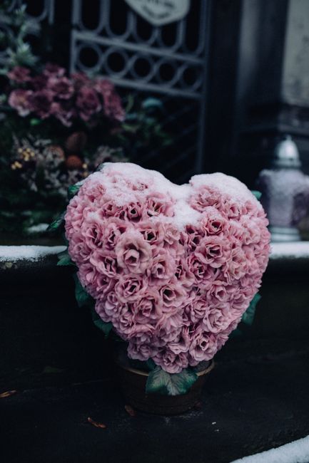 A heart-shaped floral arrangement composed of tightly clustered light pink roses, creating a textured and lush appearance. The bouquet is placed on a dark surface, with green leaves visible at the bas